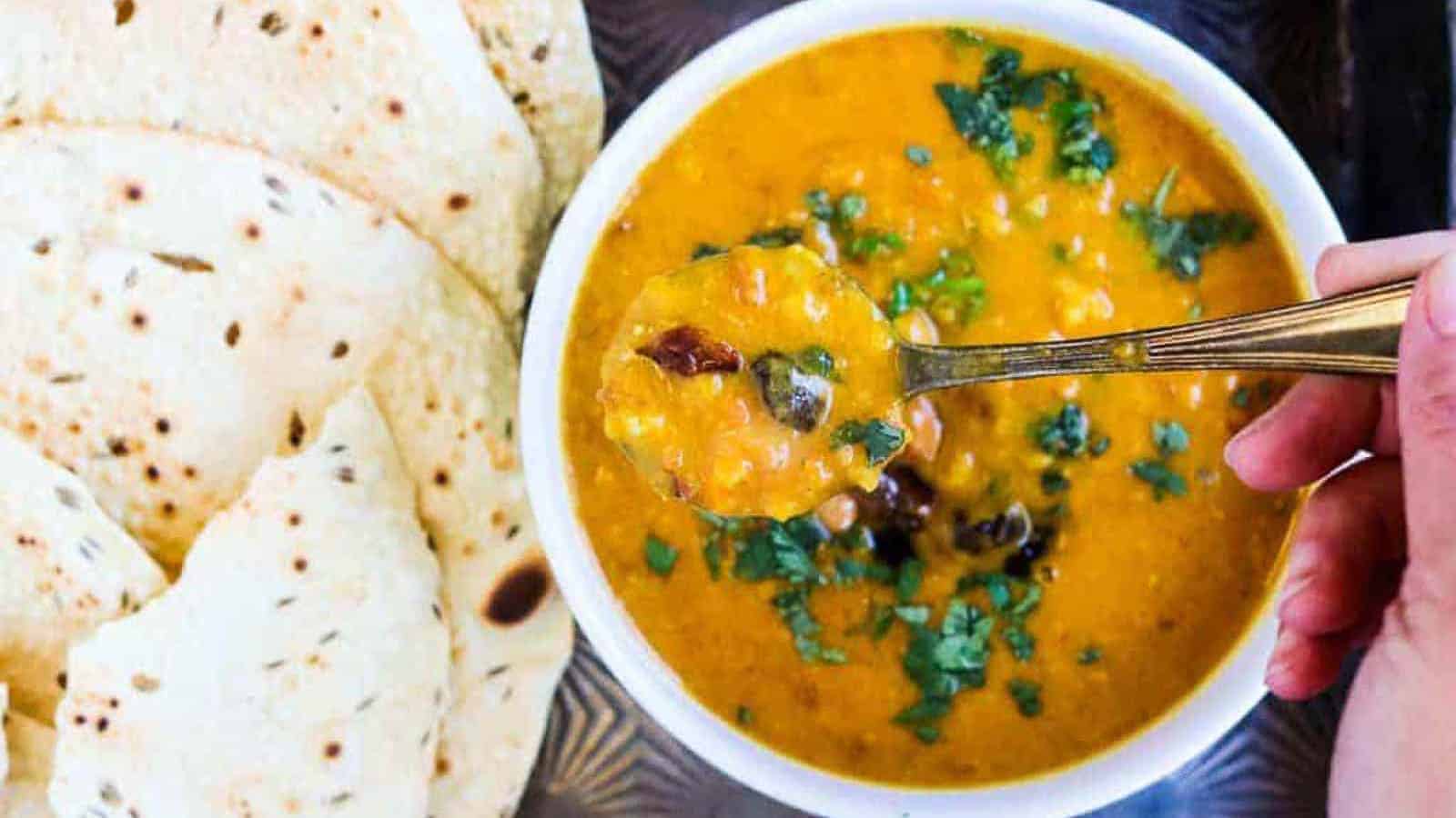 A bowl of yellow lentil dal garnished with cilantro, with a spoon and pieces of flatbread on the side.