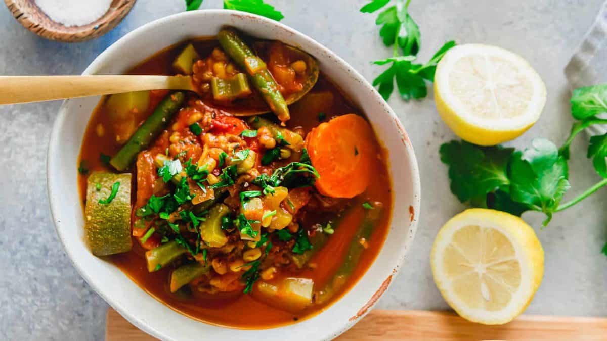 A bowl of vegetable stew with carrots, green beans, zucchini, and herbs, beside fresh parsley and two lemon halves on a gray surface.