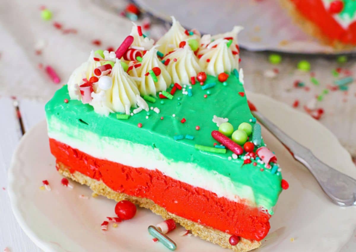 A slice of layered cheesecake with red, white, and green layers, topped with whipped cream and colorful sprinkles, on a white plate with a fork.