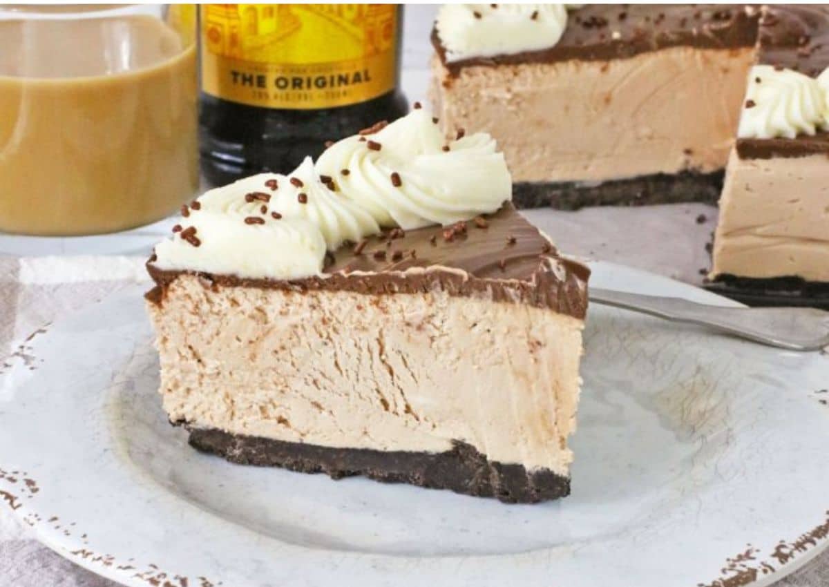 A slice of creamy cheesecake with a chocolate crust, chocolate topping, and whipped cream garnish sits on a white plate.