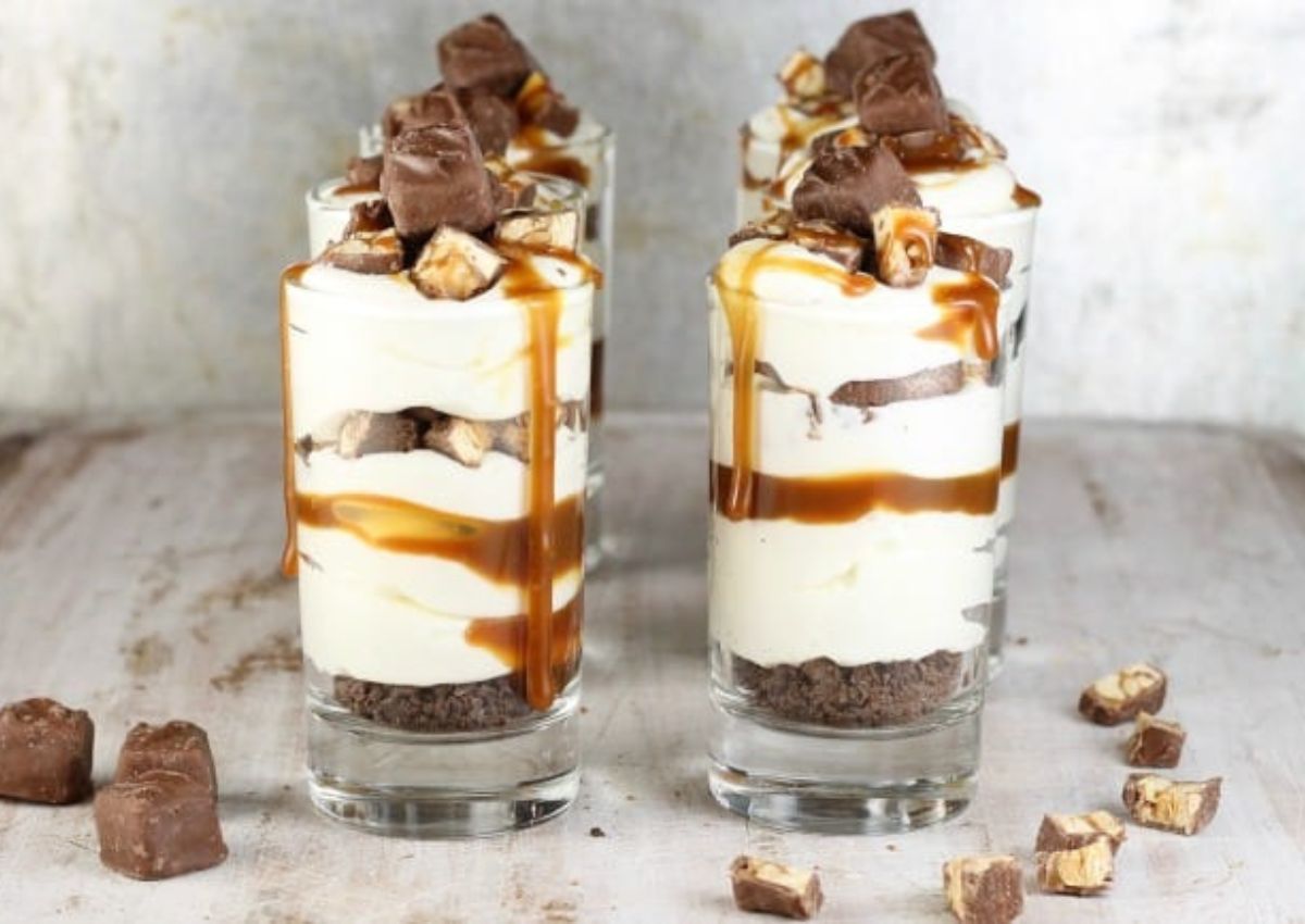 Four dessert glasses filled with layered cream, chocolate pieces, and caramel sauce, topped with chopped candy bars, sit on a light wooden surface.