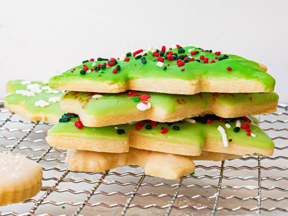 A stack of Christmas tree-shaped sugar cookies with green icing and red, white, and green sprinkles on a cooling rack.
