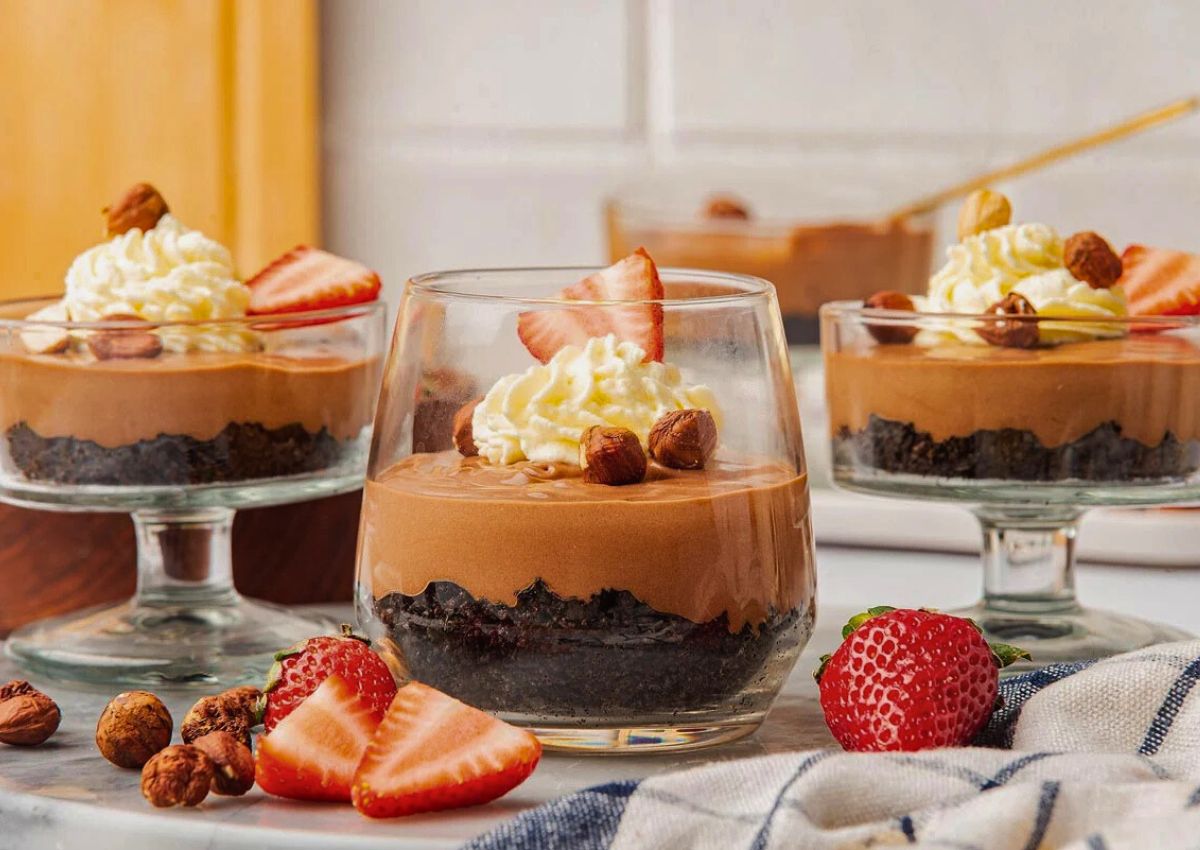 Three glass cups filled with layered chocolate mousse, crumbled cookie base, whipped cream, hazelnuts, and sliced strawberries, with extra strawberries and nuts on the table.