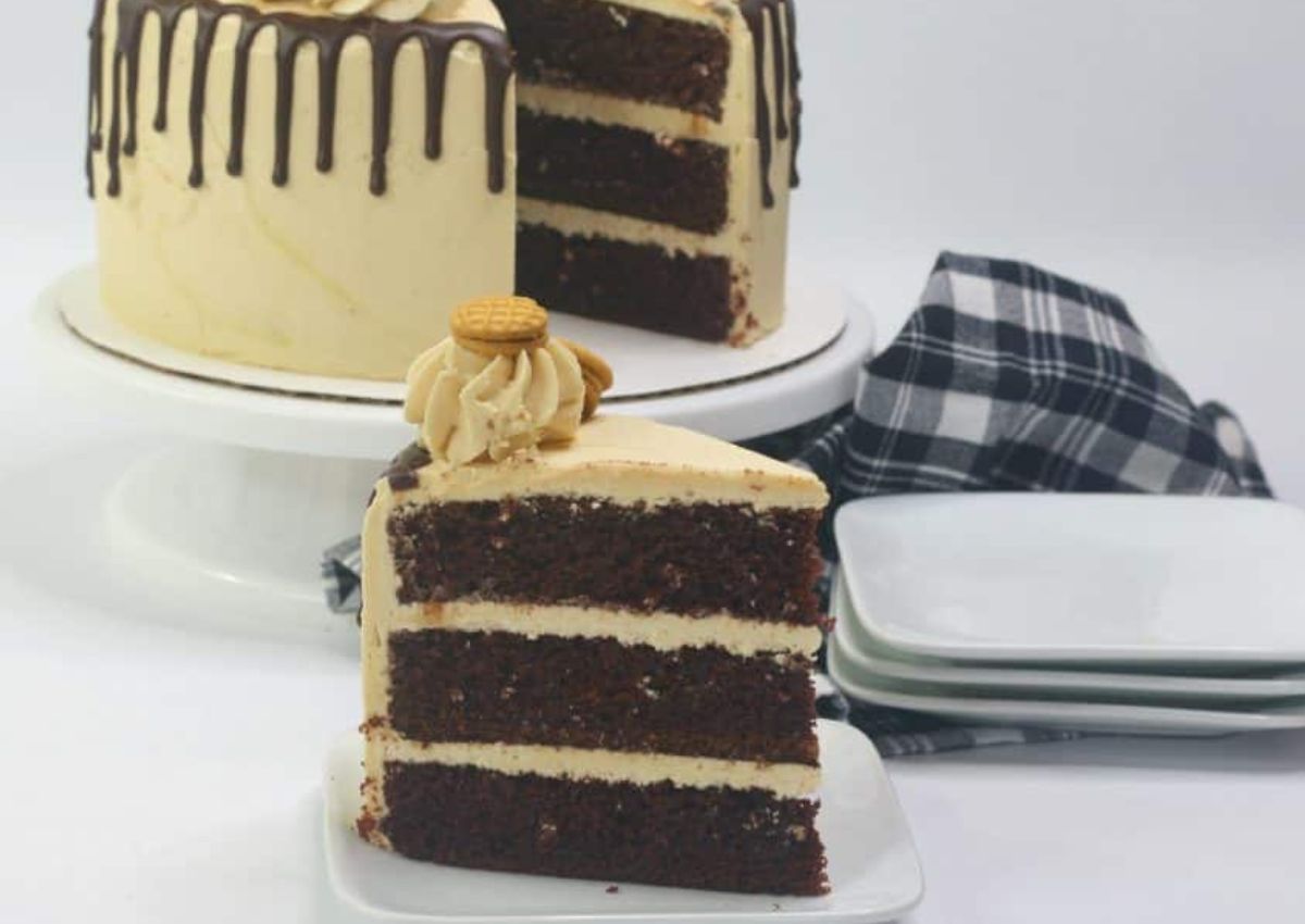 A three-layer chocolate cake with light frosting and chocolate drizzle, topped with a cookie, sits beside stacked white plates and a plaid cloth. One slice is served in the foreground.
