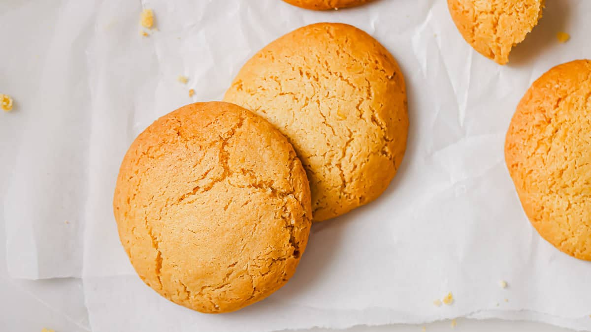 Two round, golden-brown cookies on a sheet of white parchment paper, with crumbs and a partially visible broken cookie nearby.