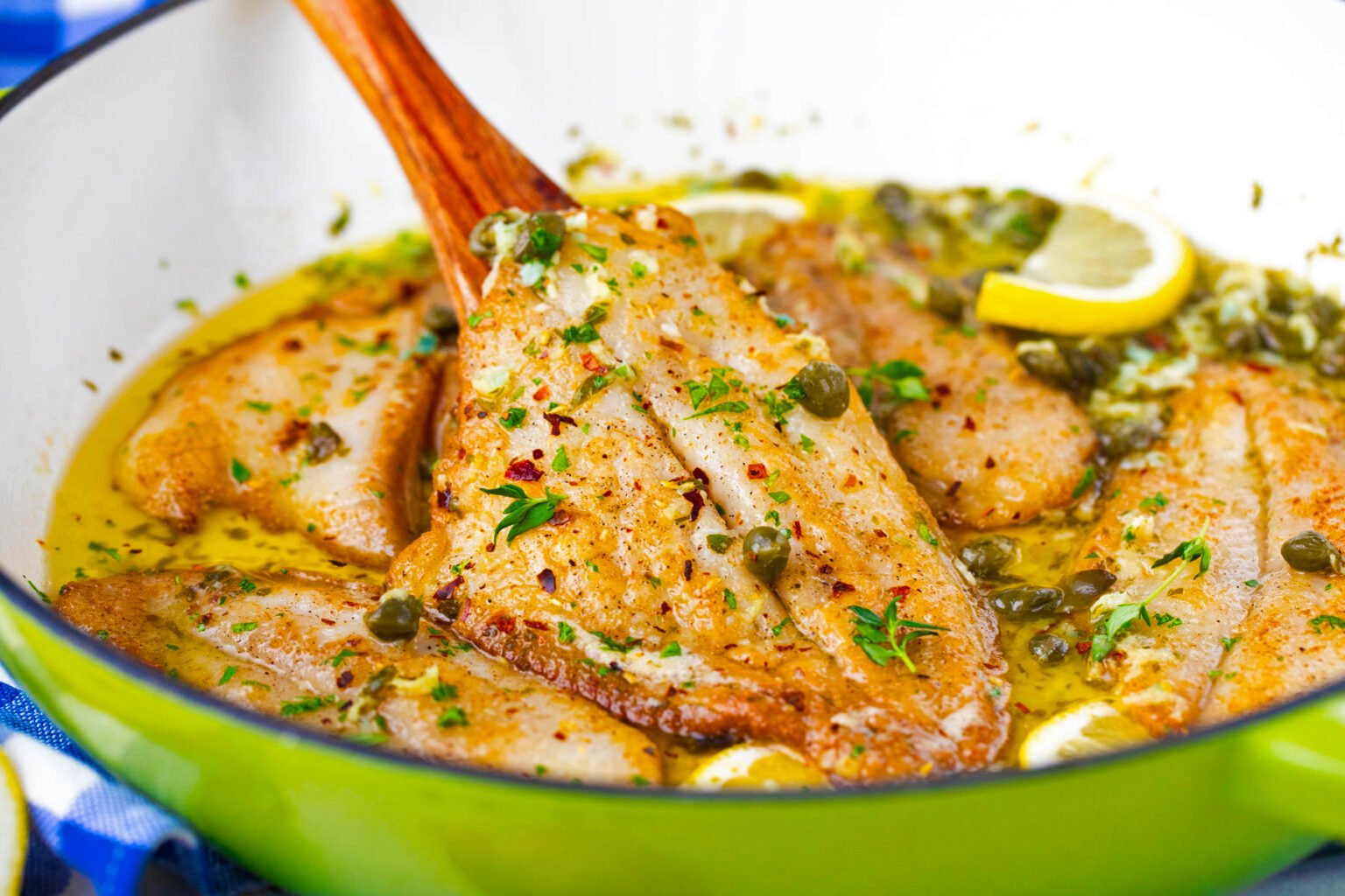 Pan-seared fish fillets in a lemon butter sauce with herbs and capers, garnished with lemon slices, in a green skillet with a wooden spatula.