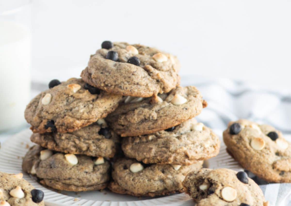 A stack of chocolate chip cookies with white and dark chocolate chips on a white plate, next to a glass of milk.