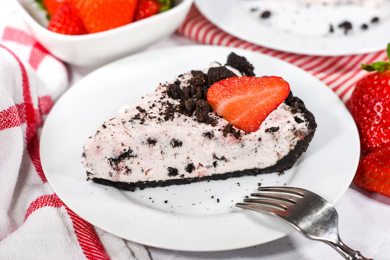A slice of cookies and cream pie with a chocolate crust, topped with crushed cookies and a sliced strawberry, served on a white plate with a fork.