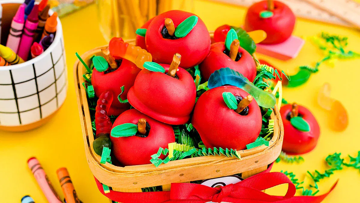 A basket of red apple-shaped treats with green leaves, decorated with gummy worms, sits on a yellow surface near crayons and pencils.