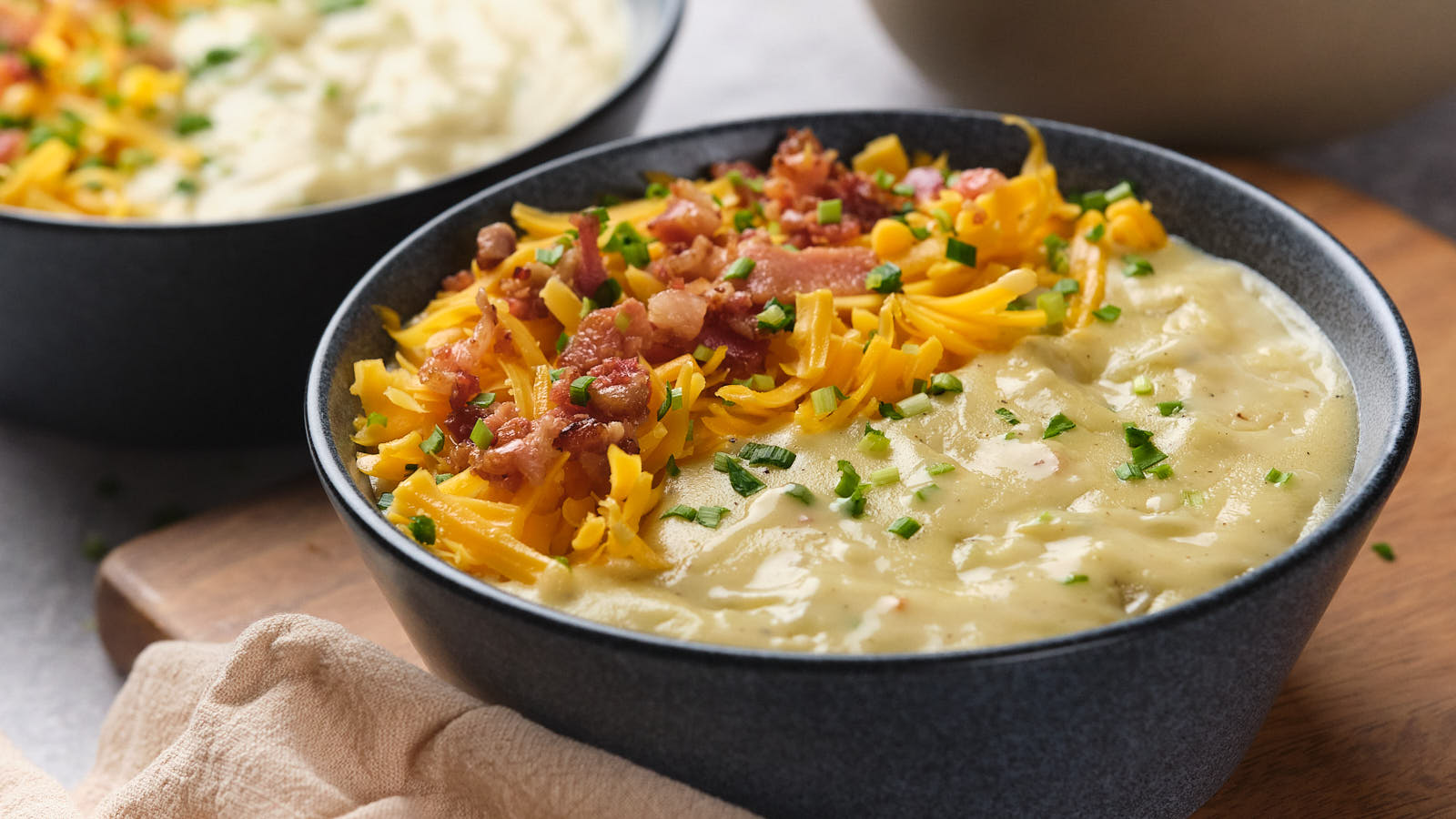 A bowl of creamy soup topped with shredded cheddar cheese, bacon bits, and chopped chives sits on a wooden surface next to a napkin.
