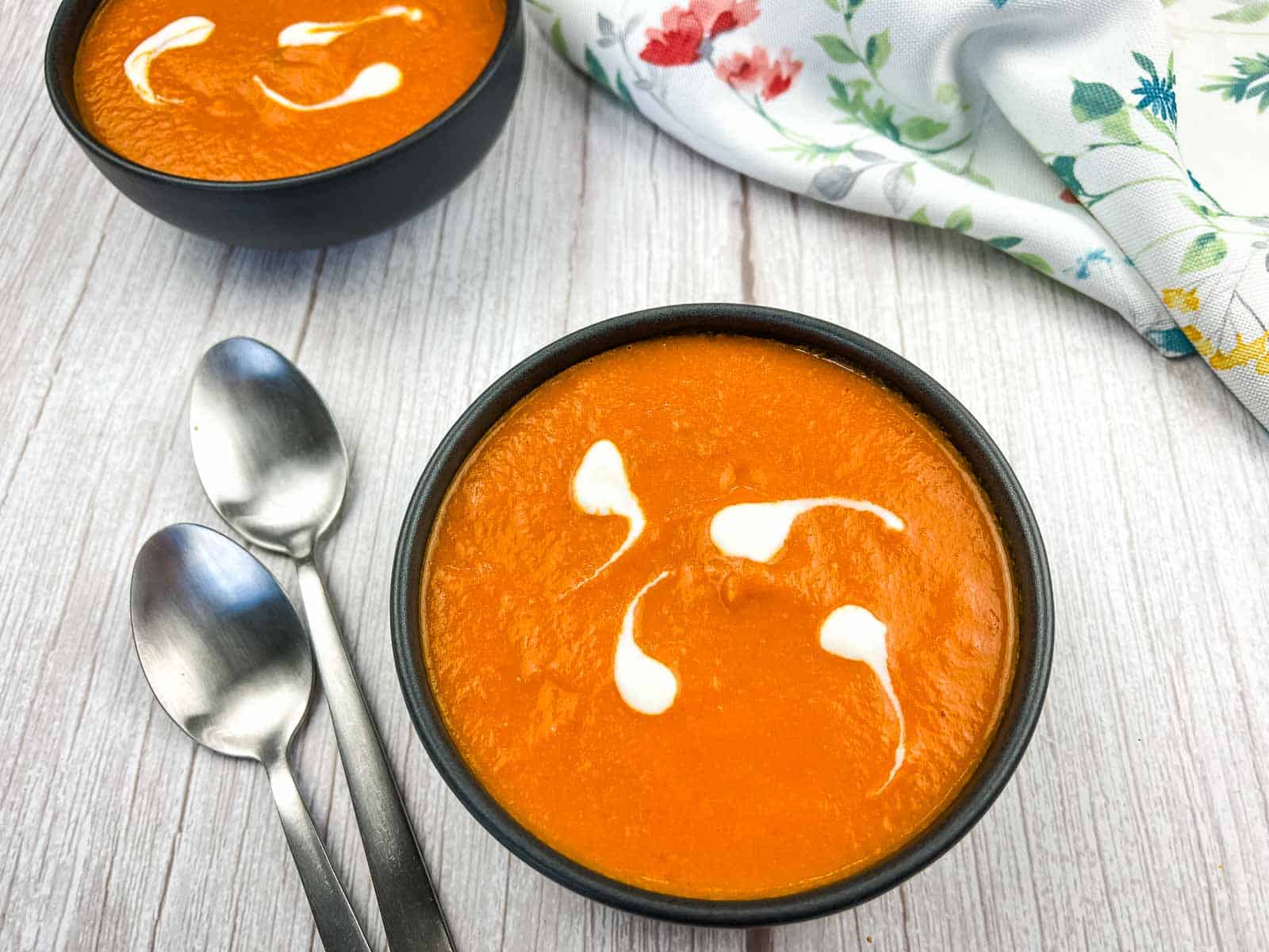 Two bowls of tomato soup with cream swirl patterns, placed on a light wooden surface next to two spoons and a floral napkin.