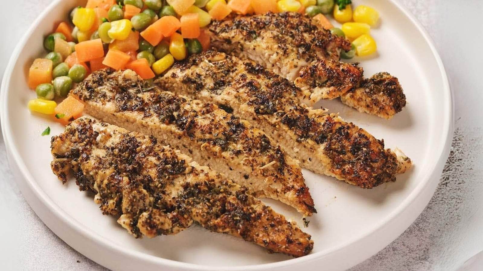 A plate with sliced, herb-seasoned grilled chicken breast and a colorful side of mixed vegetables including carrots, corn, and edamame.
