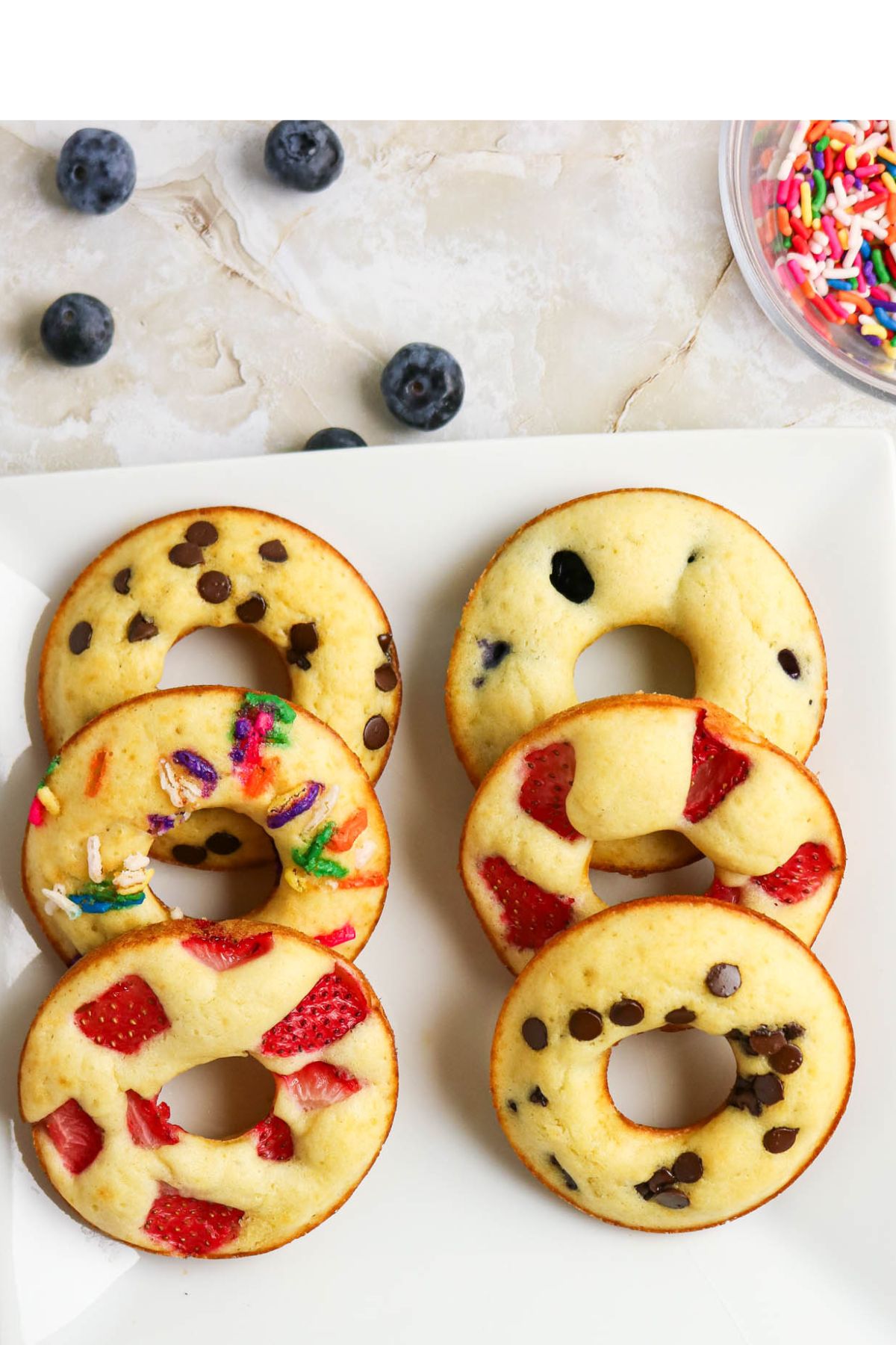 A white plate with six baked donuts, each topped with different toppings: chocolate chips, blueberries, strawberries, and colorful sprinkles. Blueberries and sprinkles are nearby.