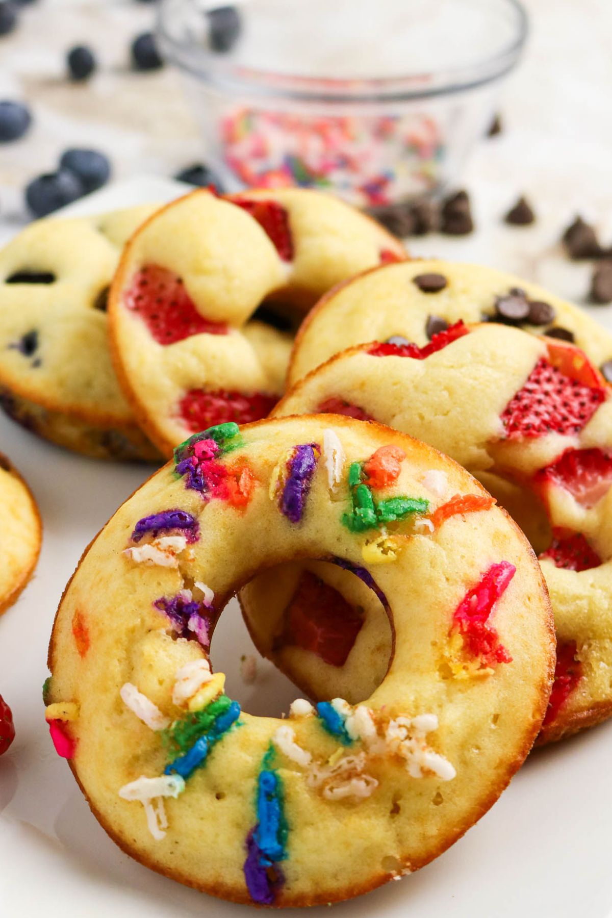 A close-up of baked donut-shaped treats topped with colorful sprinkles, strawberries, and chocolate chips on a white plate, with a blurred bowl in the background.