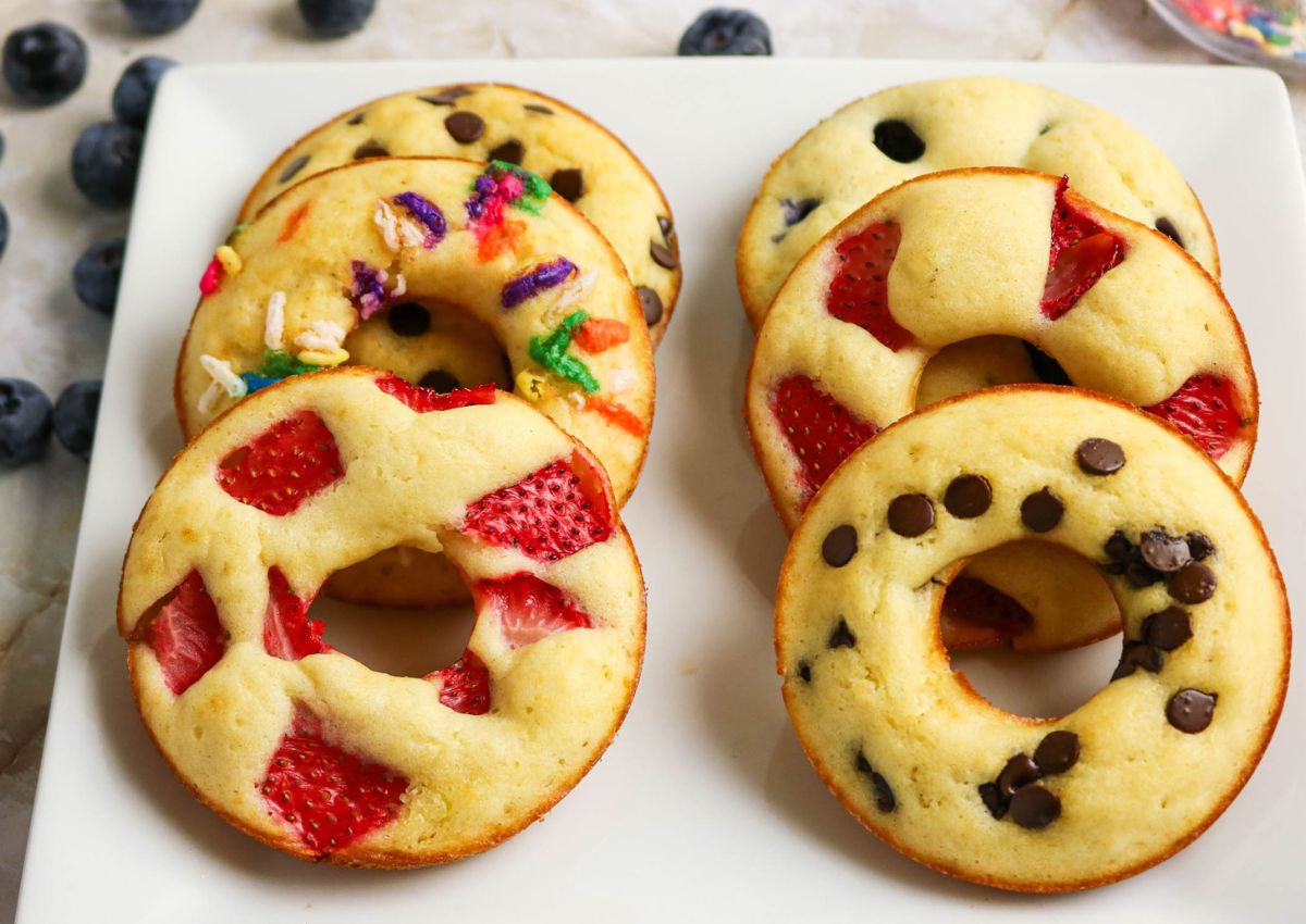 Six baked donuts on a white plate, each topped with different mix-ins including sprinkles, strawberries, and chocolate chips. Blueberries are scattered in the background.