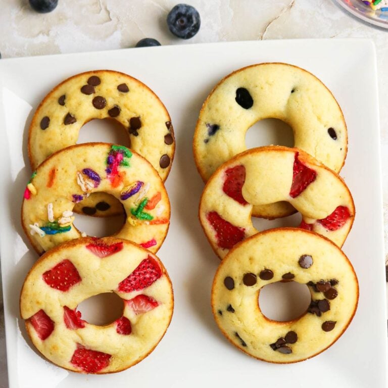 A white plate with six baked donuts topped with chocolate chips, sprinkles, blueberries, and strawberries, arranged in two rows. Blueberries are scattered nearby.