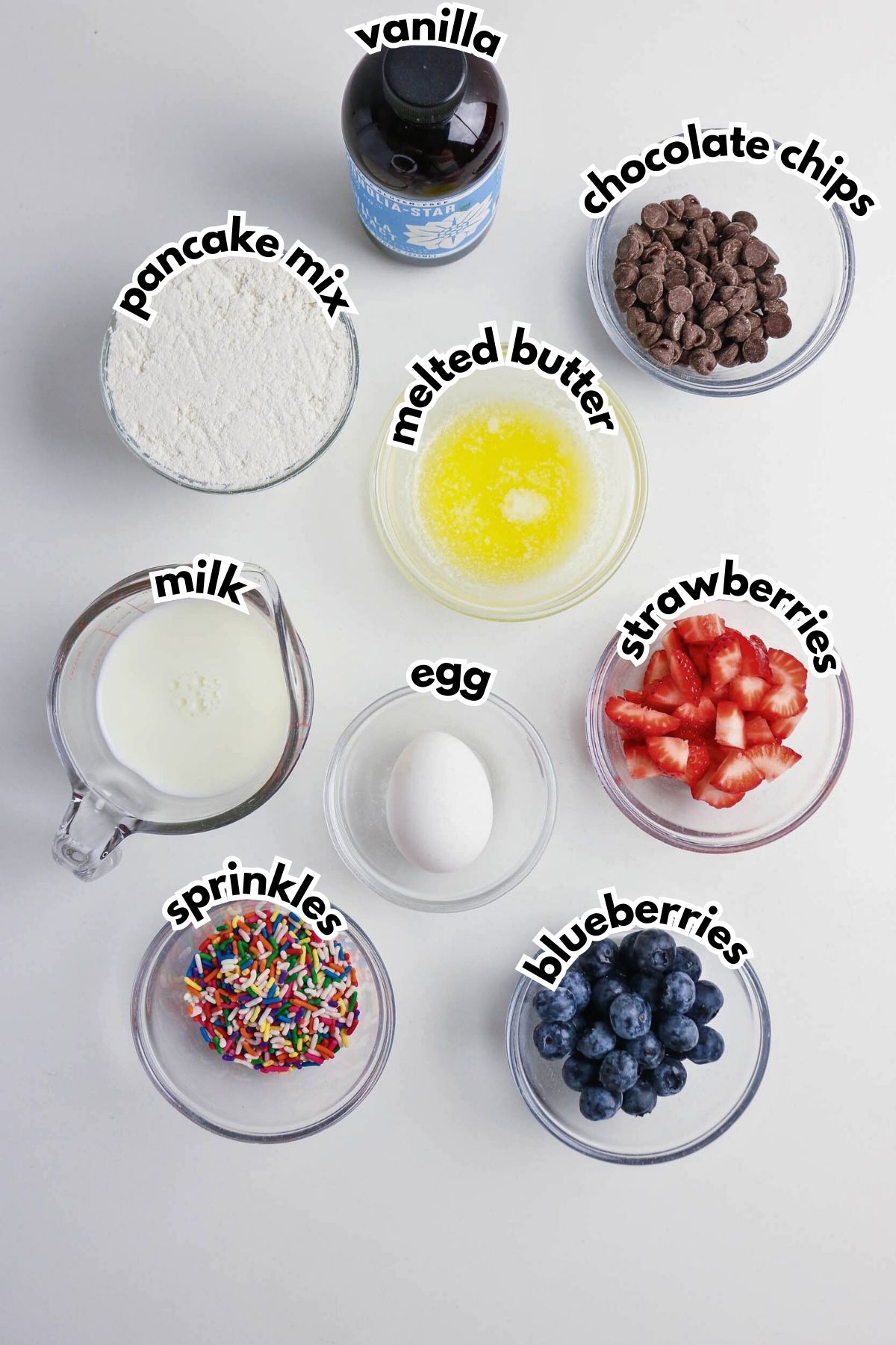 Top-down view of ingredients in bowls and cups: pancake mix, chocolate chips, melted butter, vanilla, milk, egg, chopped strawberries, blueberries, and sprinkles.