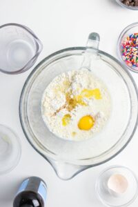 A glass mixing bowl with flour, sugar, milk, egg, and melted butter sits on a white surface surrounded by measuring cups, sprinkles, vanilla extract, and an eggshell.