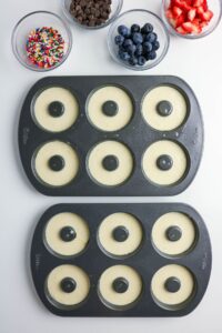 Two donut pans filled with batter are on a white surface, with bowls of sprinkles, chocolate chips, blueberries, and chopped strawberries beside them.