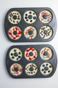 Two donut pans filled with batter, each donut topped with different ingredients including chocolate chips, blueberries, strawberries, and colorful sprinkles, on a white surface.