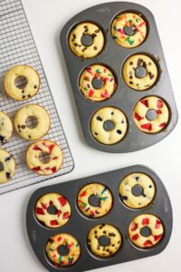 Three donut pans filled with baked donuts topped with chocolate chips, sprinkles, and fruit. A cooling rack holds several finished donuts.