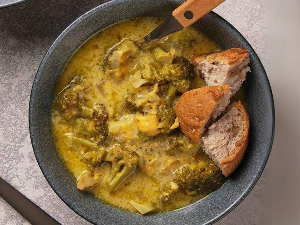 A bowl of broccoli and cheese soup is served with two pieces of bread; a spoon rests on the edge of the bowl.