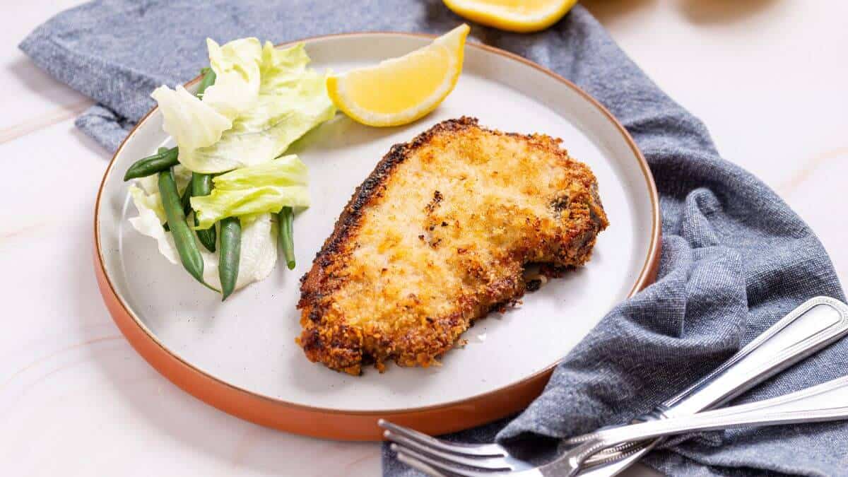 A breaded and fried pork chop served with a lemon wedge, lettuce, and green beans on a round plate with a fork and knife beside it.