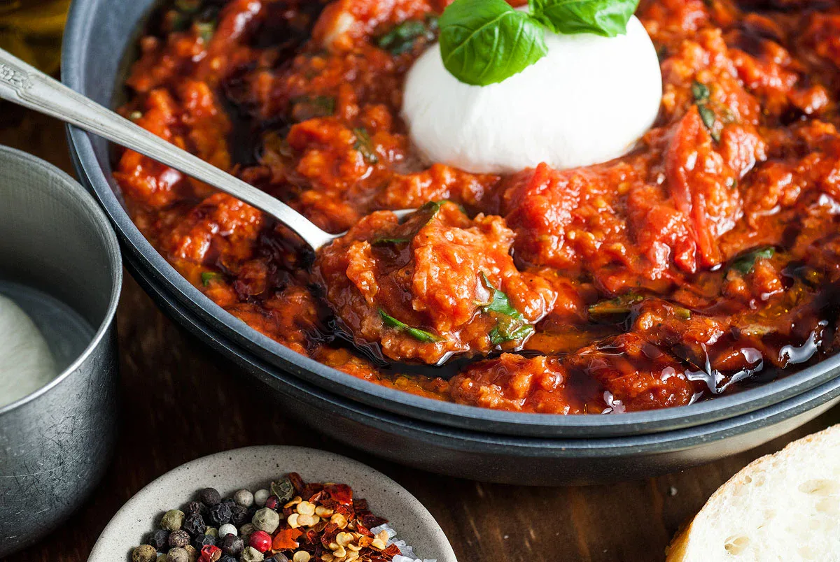 A bowl of tomato-based stew topped with a ball of mozzarella and fresh basil, with a spoon, a bowl of spices, and a slice of bread nearby.