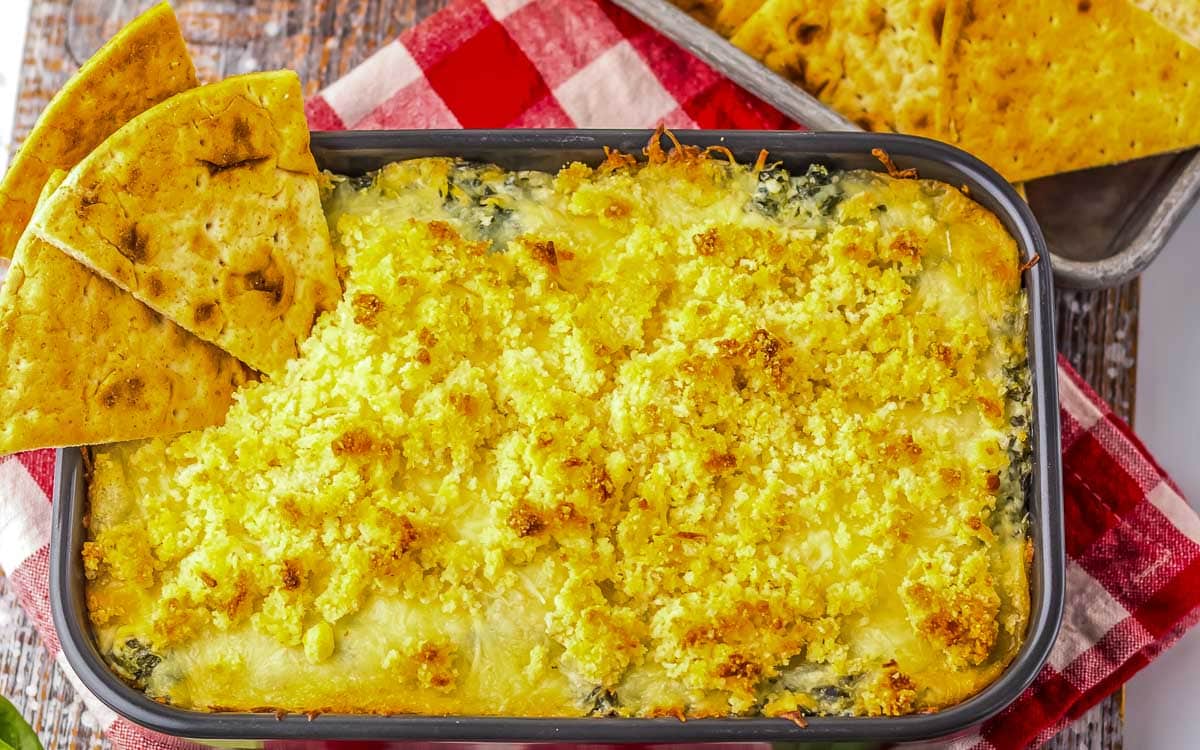 Baking dish filled with cheesy spinach and artichoke dip topped with breadcrumbs, served with triangular pita chips and rectangular crackers on a red and white checkered cloth.