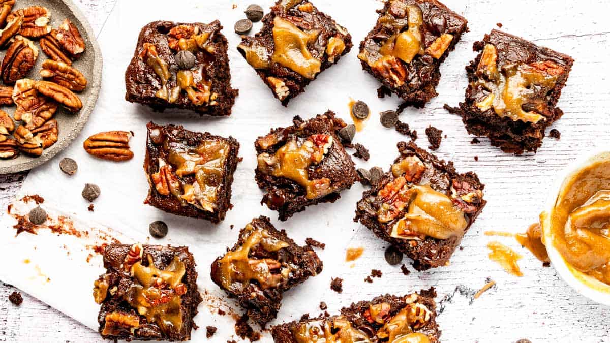 Nine chocolate pecan brownies topped with caramel are arranged on a white surface, with scattered pecans, chocolate chips, and a bowl of caramel sauce nearby.