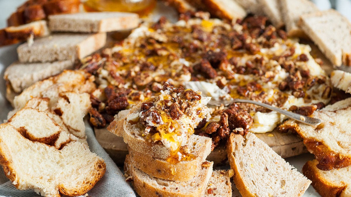 A platter of sliced bread surrounds a cheese spread topped with nuts and honey, with a serving spoon resting on top.