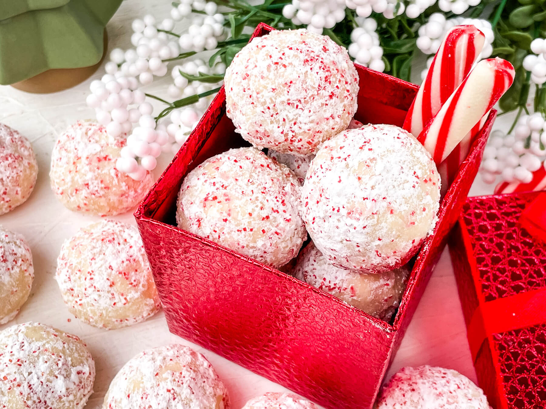 A red box filled with powdered sugar-coated cookies and two candy canes, with more cookies and decorative white berries in the background.