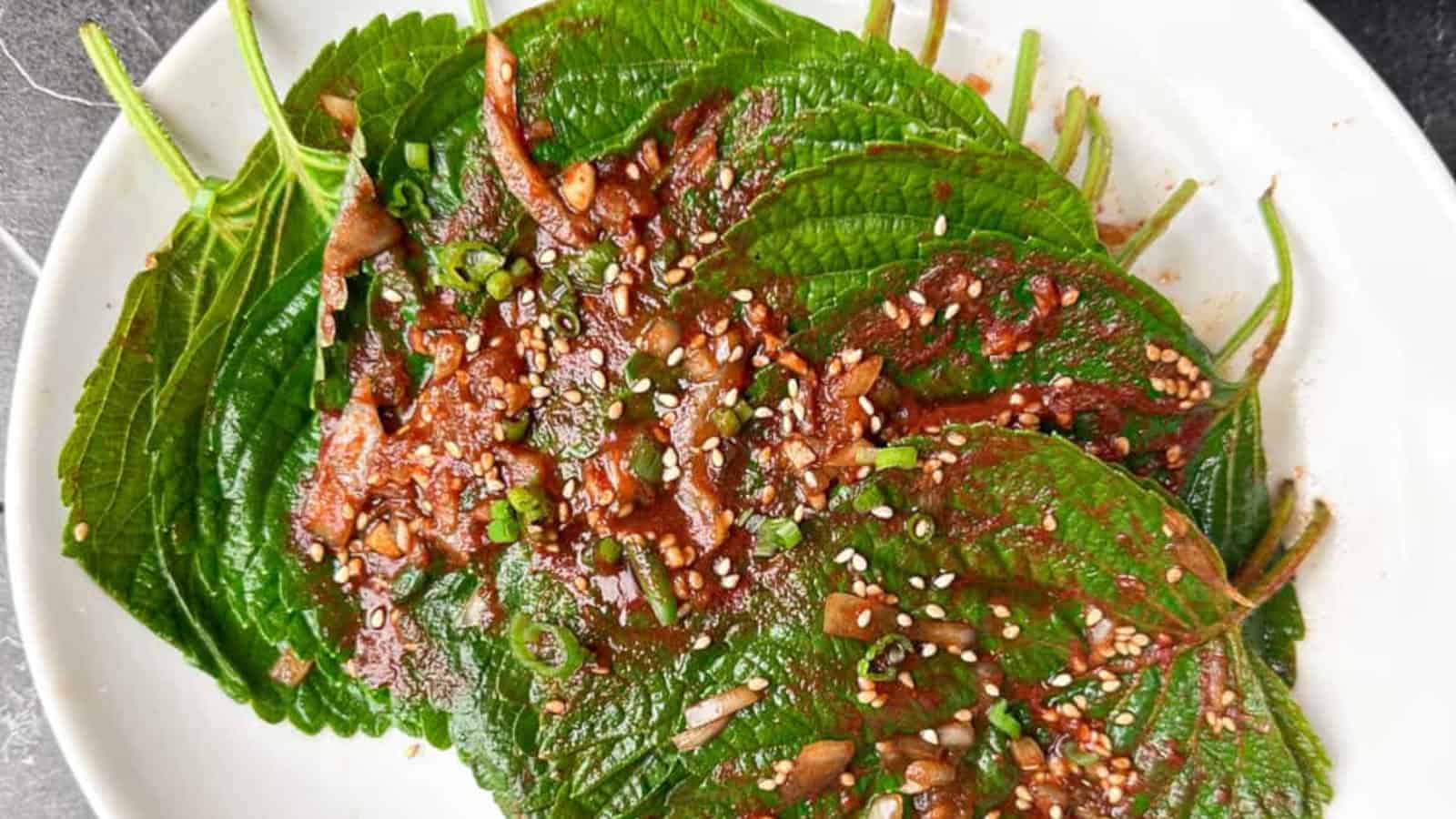 A plate of green perilla leaves topped with chopped garlic, green onions, sesame seeds, and a red chili paste seasoning.