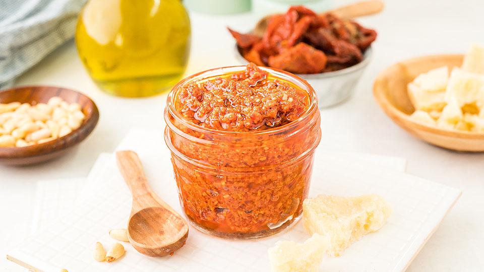 A small glass jar filled with red pesto sits on a white plate, surrounded by pine nuts, sun-dried tomatoes, olive oil, Parmesan cheese, and a wooden spoon.
