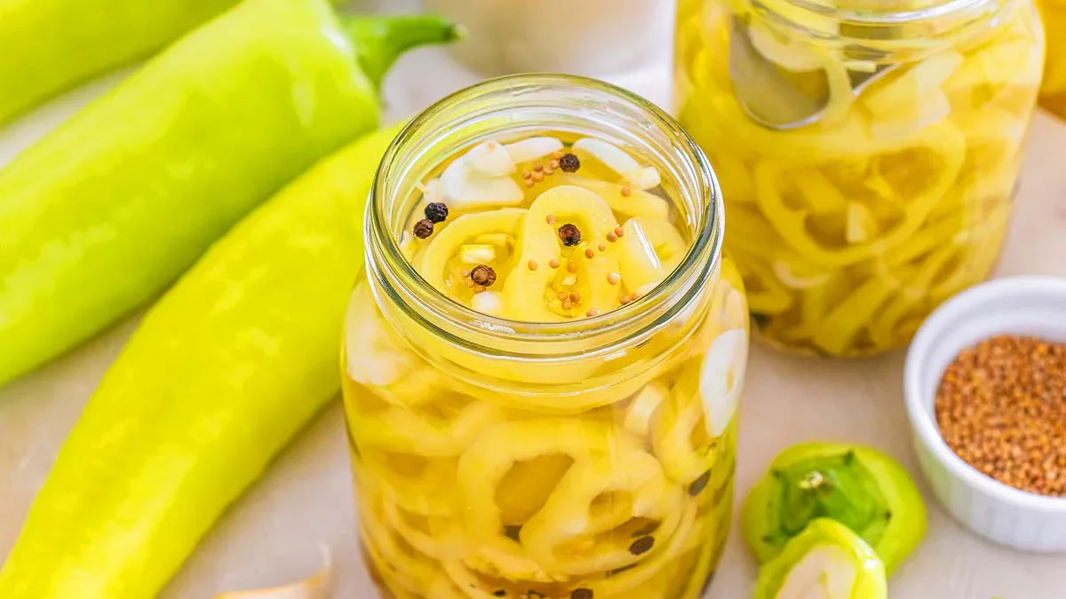 Two jars filled with pickled banana pepper rings, surrounded by fresh banana peppers and pickling spices on a light surface.