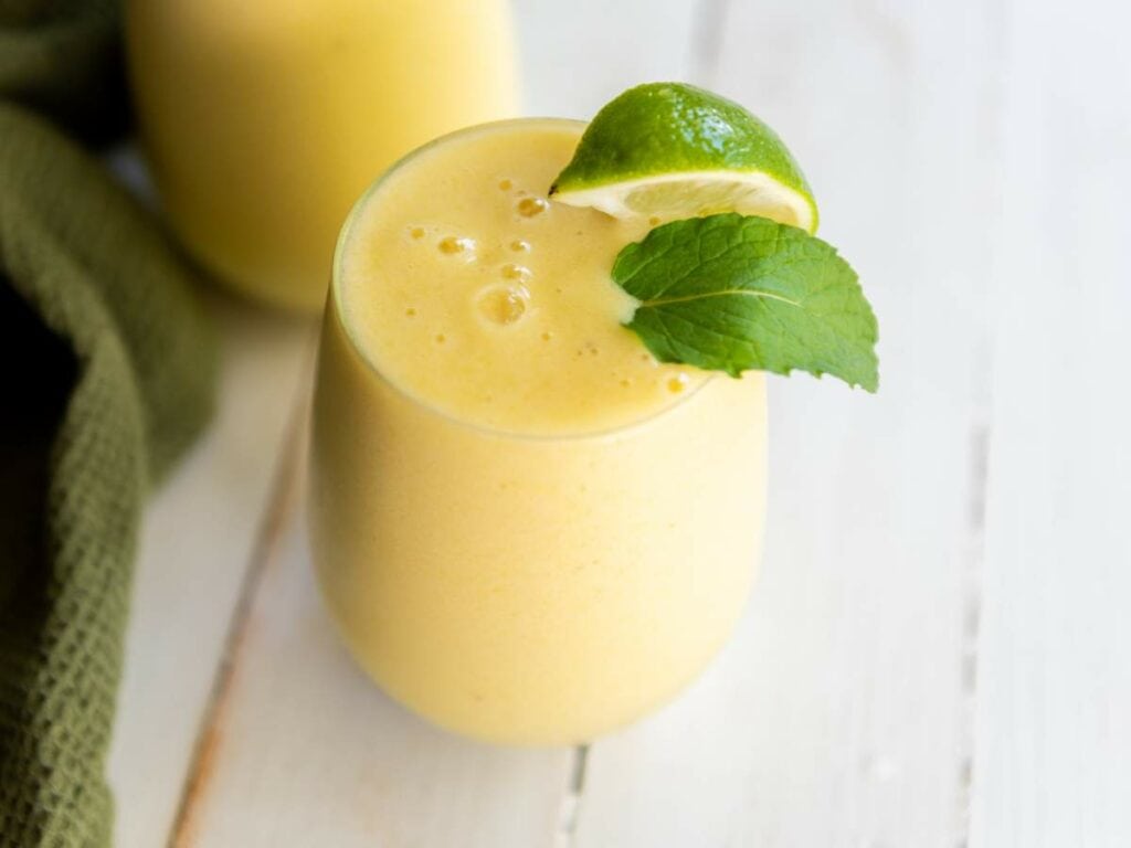 A glass of yellow smoothie garnished with a lime wedge and a mint leaf, placed on a white wooden surface.