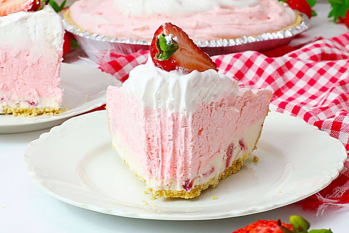 A slice of pink strawberry cream pie with a graham cracker crust, topped with whipped cream and a fresh strawberry, on a white plate.