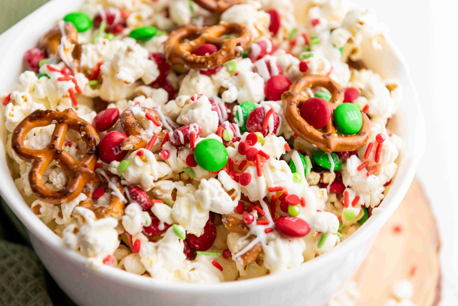 A bowl filled with popcorn, pretzels, red and green chocolate candies, and festive sprinkles, drizzled with white icing.