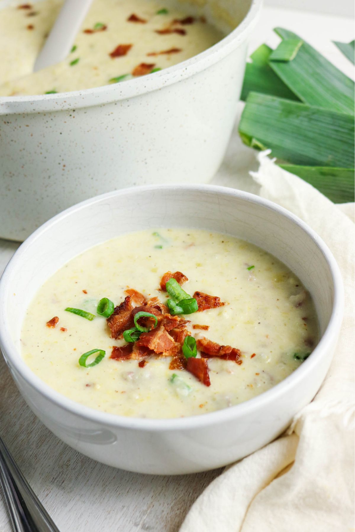 A bowl of creamy soup topped with chopped bacon and green onions, with a pot of soup and fresh leeks in the background.