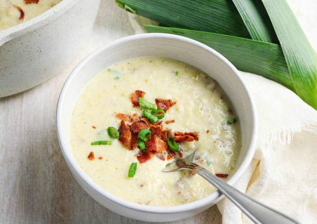 A bowl of creamy soup topped with chopped bacon and green onions, with a spoon in the bowl and fresh leeks and a napkin beside it.