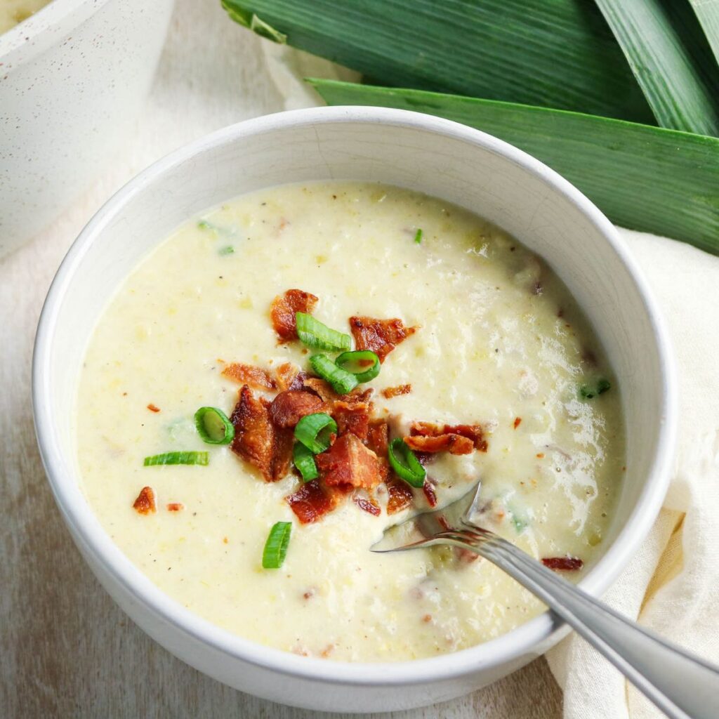 A bowl of creamy soup topped with crumbled bacon and chopped green onions, with a spoon resting inside the bowl.