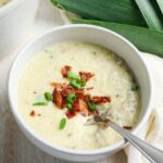 A bowl of creamy soup topped with crumbled bacon and chopped green onions, with a spoon resting inside the bowl.