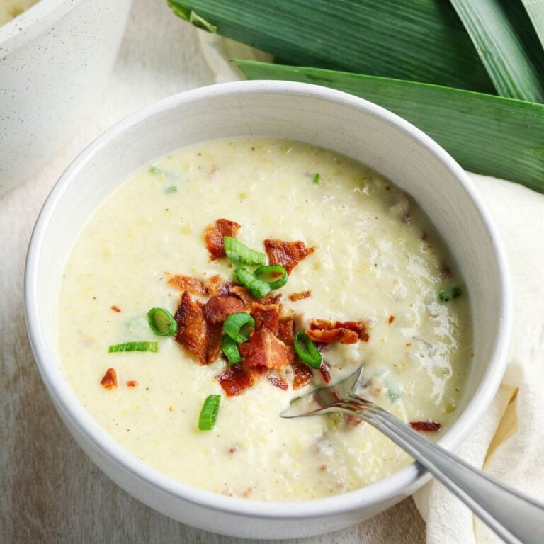 A bowl of creamy soup topped with crumbled bacon and chopped green onions, with a spoon resting inside the bowl.