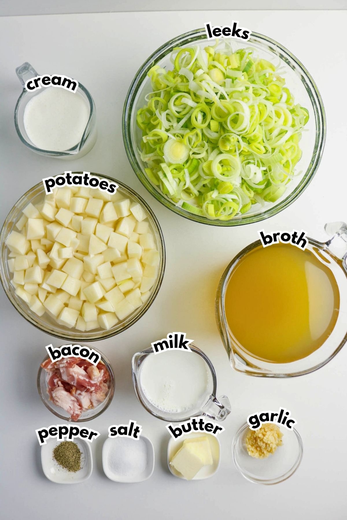 Ingredients for leek and potato soup arranged on a white surface, including cream, leeks, potatoes, broth, bacon, milk, butter, garlic, salt, and pepper in separate bowls.