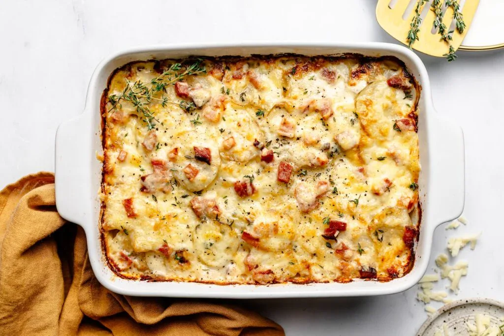 A baked casserole topped with melted cheese and diced ham, garnished with fresh thyme in a white rectangular dish on a white surface.