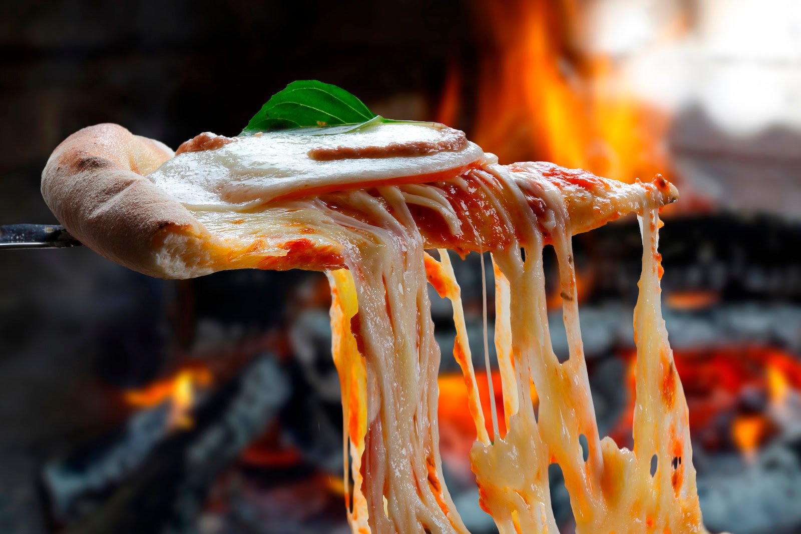 A slice of pizza with melted cheese and basil is lifted in front of a wood-fired oven with visible flames in the background.
