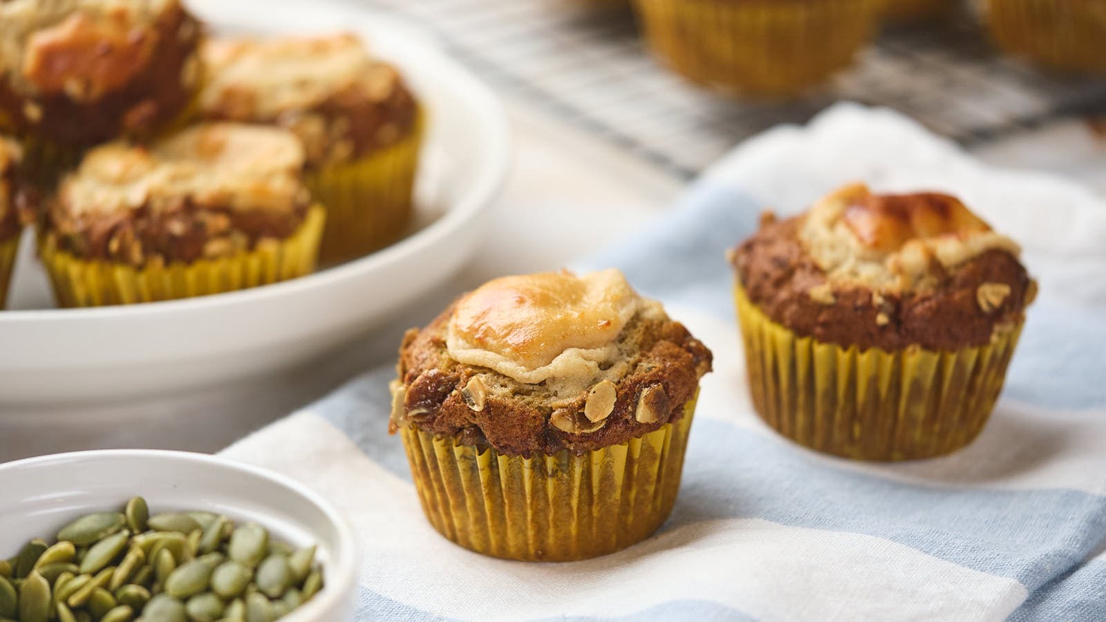 Two pumpkin muffins with cream cheese swirls in yellow paper liners rest on a blue and white cloth, with more muffins and a bowl of pumpkin seeds nearby.