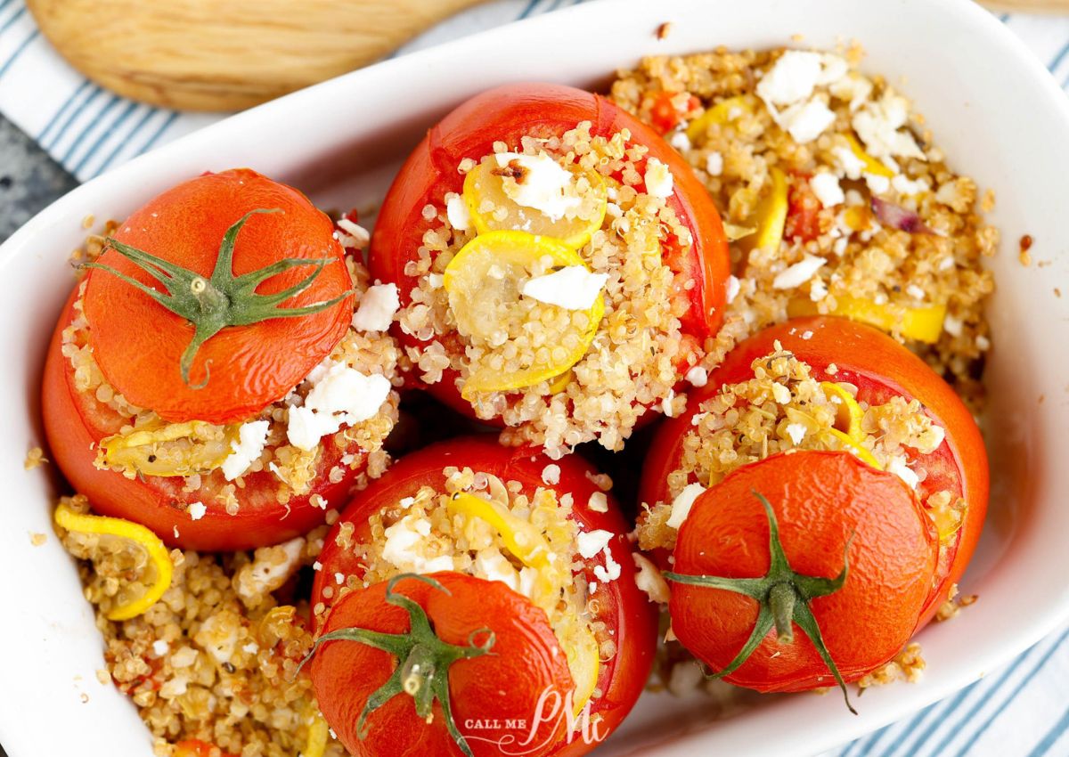 Four baked tomatoes stuffed with quinoa, yellow bell pepper slices, and crumbled feta cheese, arranged in a white dish.
