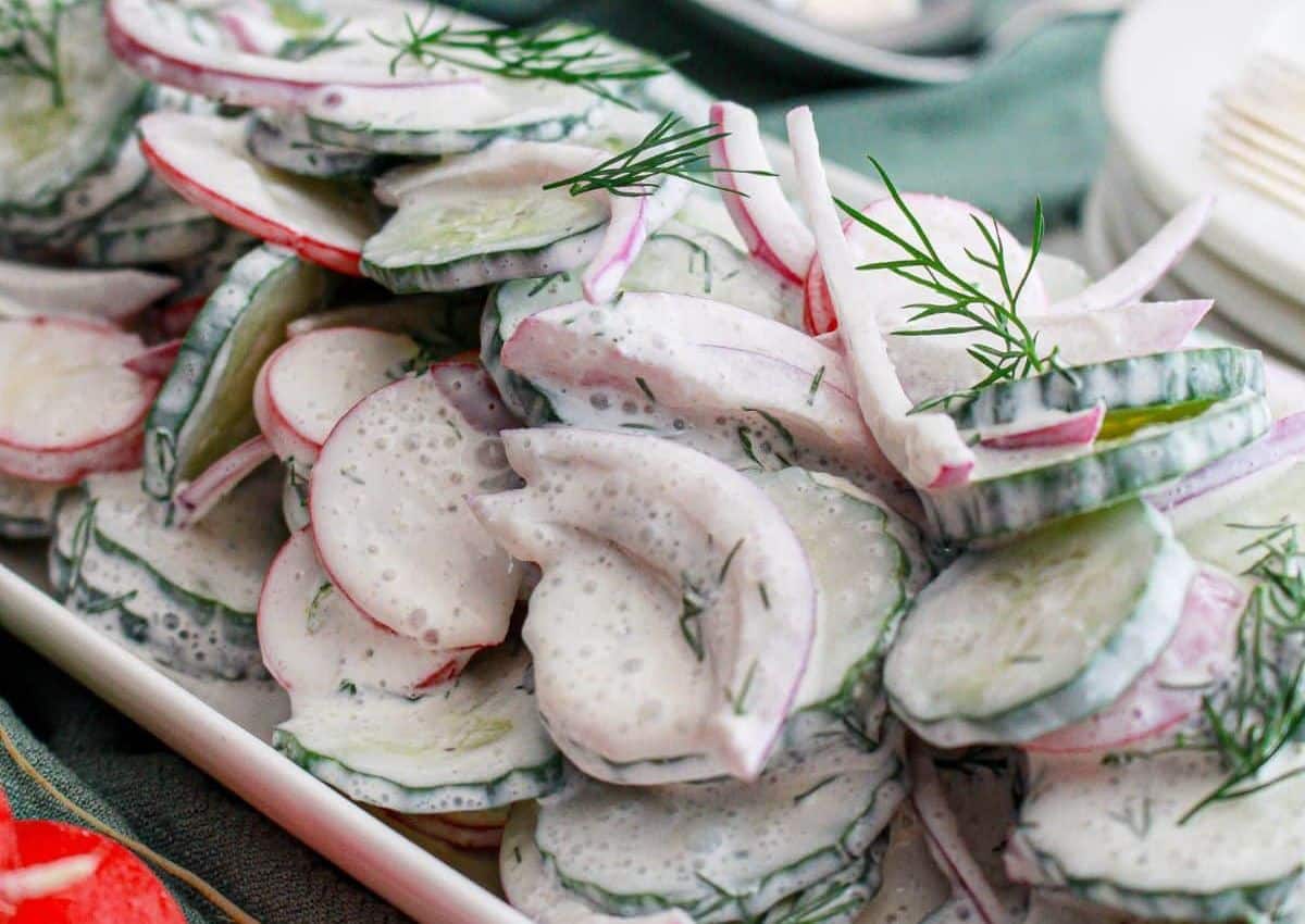 Sliced cucumbers, radishes, and red onions coated in a creamy dressing, garnished with fresh dill on a white plate.