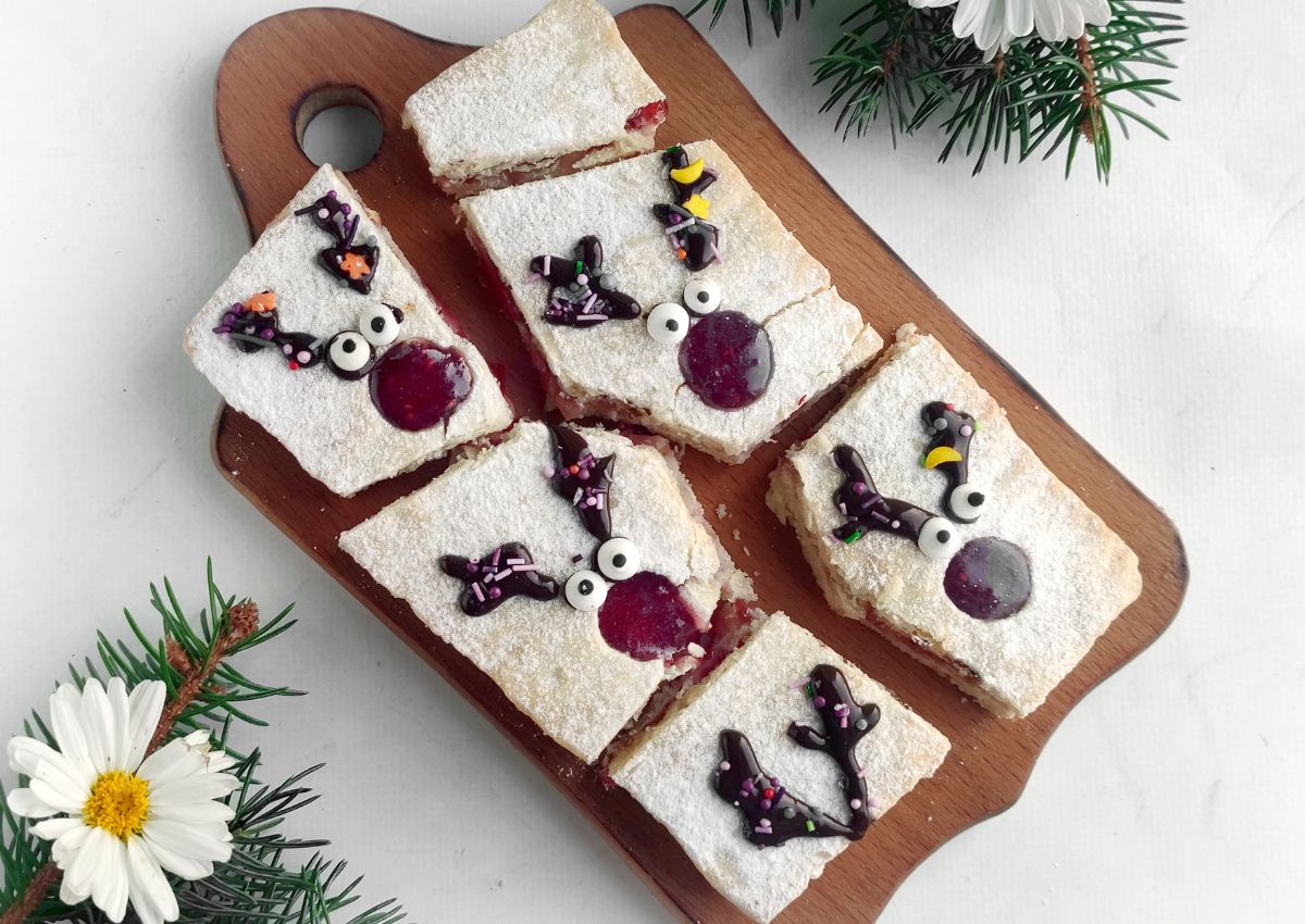 Six rectangular pastries dusted with powdered sugar, decorated with icing and candy to resemble reindeer faces, are arranged on a wooden serving board. Pine branches and a daisy are nearby.